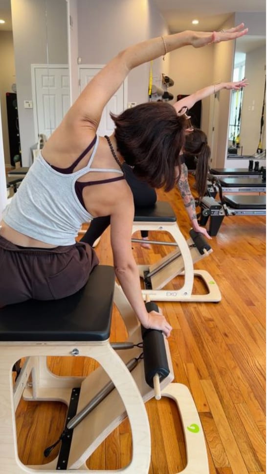 Two clients seen from behind on Pilates chairs, each reaching one arm overhead in a side bend