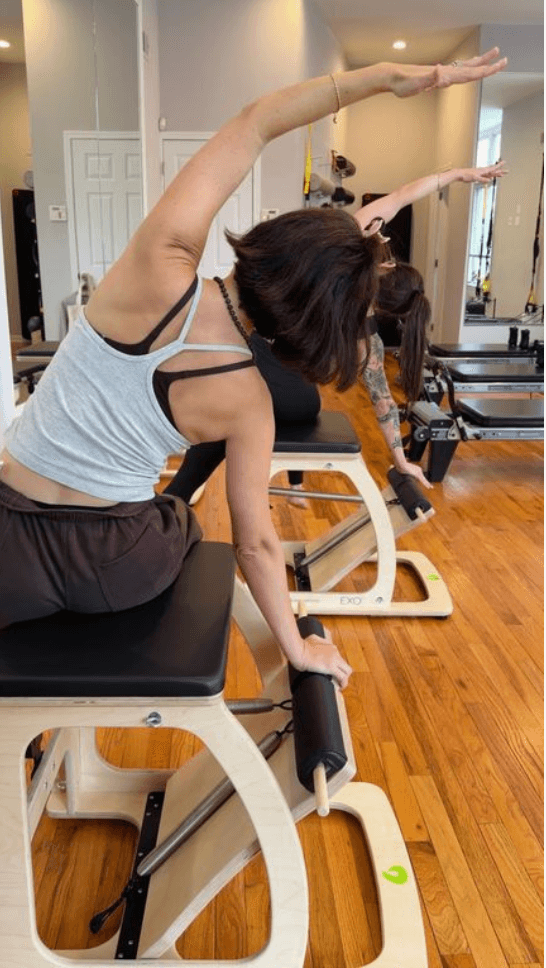 Two clients seen from behind on Pilates chairs, each reaching one arm overhead in a side bend